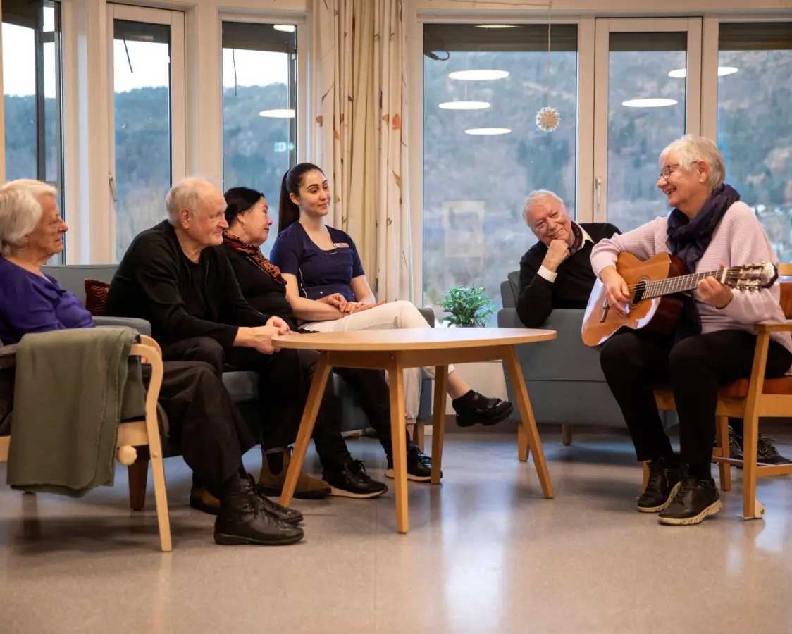 En kvinne underholder med gitar på stuen på et sykehjem. Publikum er beboere og en ansatt