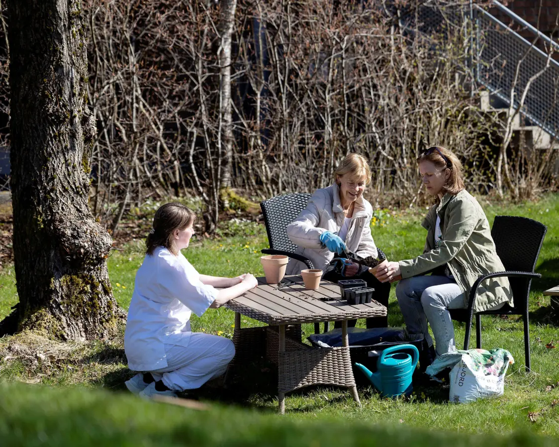 En eldre og en ung kvinne sitter i hagen ved et bord og jobber med planter sammen med en sykepleier.