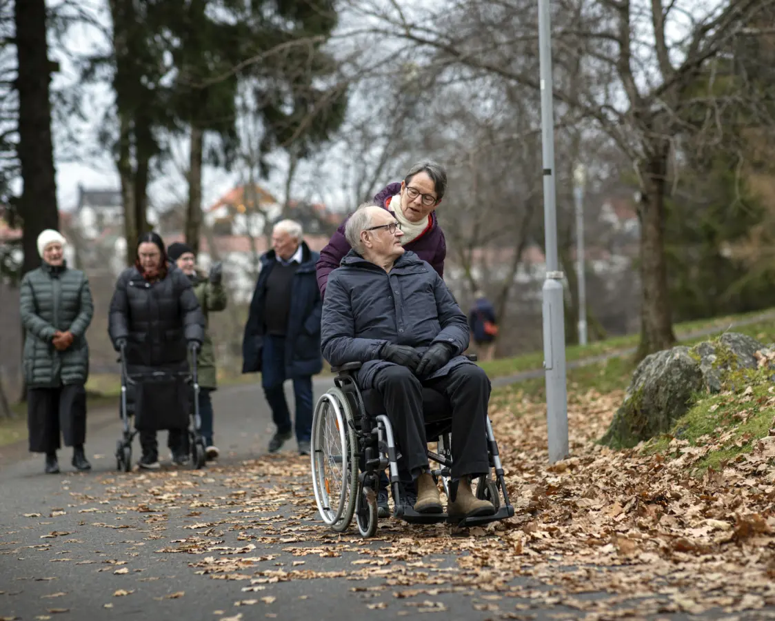 En gruppe eldre er ute på tur. I forgrunnen ser vi en mann som sitter i rullestolen sammen med en kvinne