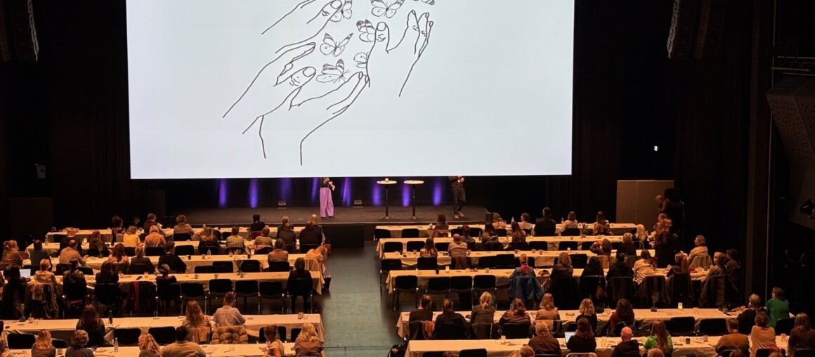 To personer sitter rundt et bord og hører på en som forteller og viser på en tavle