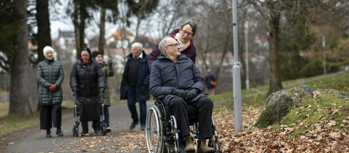 En gruppe eldre er ute på tur. I forgrunnen ser vi en mann i rullestol, og en kvinne som triller rullestolen