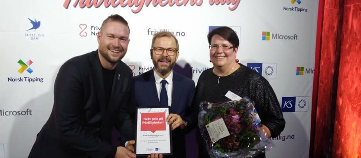To menn og en kvinne står foran et banner hvor det står Frivillighetens dag