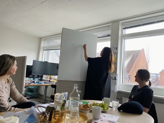 To personer sitter rundt et bord, og en underviser