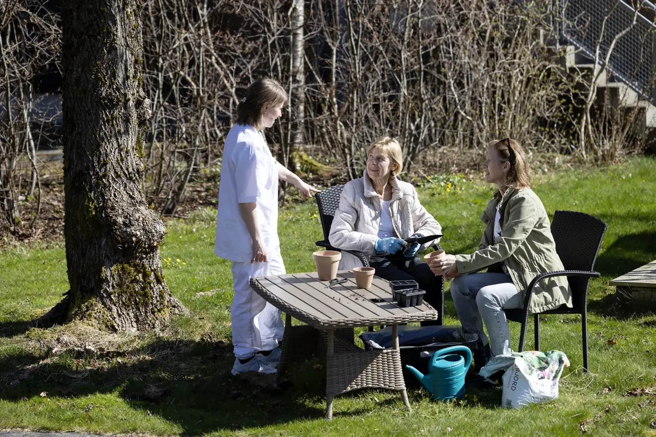 En eldre kvinne, en ung frivillig og en kvinnelig sykepleier snakker sammen ute i hagen.