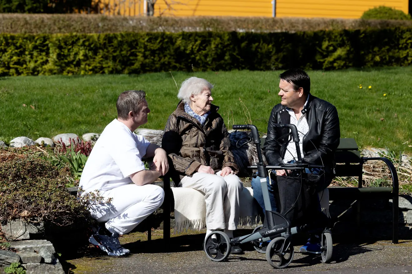 En eldre kvinne sitter på en benk sammen med to menn, en frivillig og en ansatt